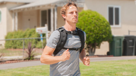 Man jogging outdoors wearing a weighted vest, with houses and greenery in the background.
