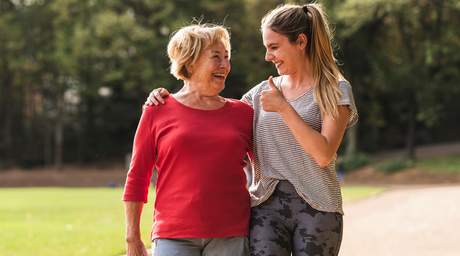 An older woman and a younger woman walking arm in arm outdoors, smiling and enjoying each other's company on a sunny day.