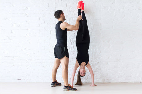 Man assisting woman with a handstand exercise against a white wall during strength and balance training.